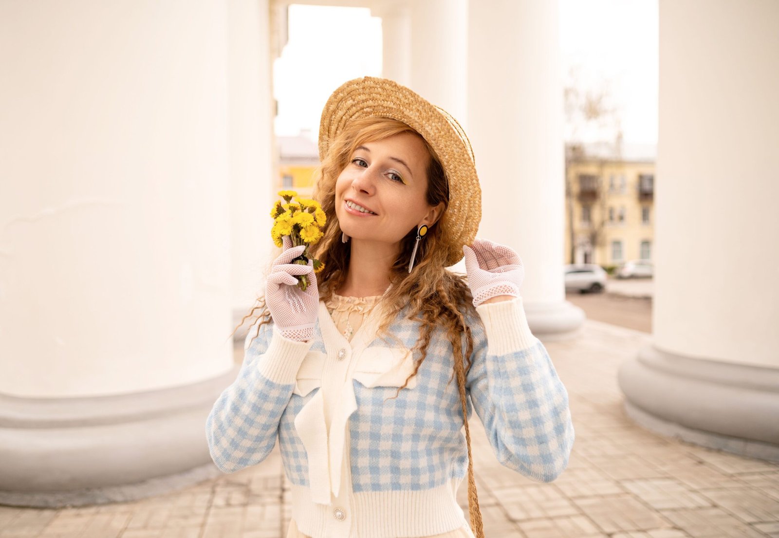 Stylish woman smiling in a hat holding yellow flowers, pillars in background.